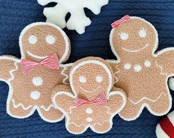 May include: Three gingerbread man-shaped cushions with white icing details and red and white striped bows. The cushions are on a blue knitted fabric.