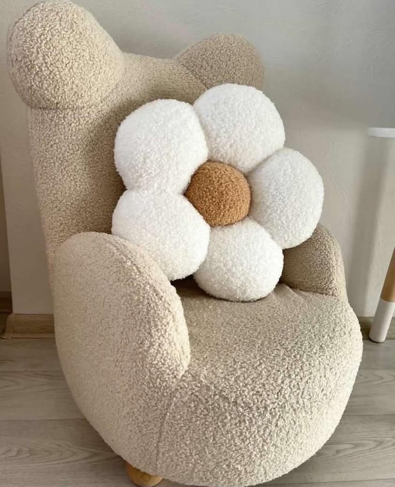 May include: A beige teddy bear-shaped armchair with a large white and brown flower-shaped pillow. The chair and pillow have a soft, textured appearance. The chair has a rounded back and armrests.