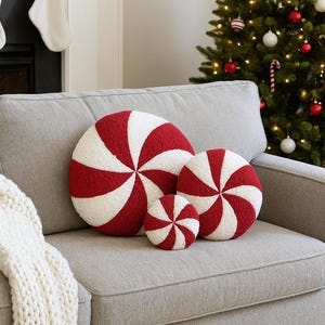 May include: A set of four round decorative pillows in red and white, resembling peppermint candies, arranged on a gray sofa. The pillows vary in size, with a large Christmas tree in the background.
