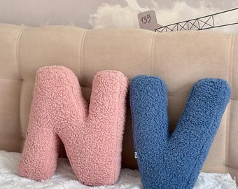 May include: Two plush letter pillows, one pink "N" and one blue "V", are sitting on a white couch.