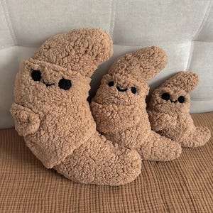 Puede incluir: Tres cojines de peluche marrones con forma de emoji de caca sonriente de varios tamaños. Cada uno tiene ojos bordados negros y una boca curvada. Los cojines están hechos de un material suave y texturizado y están colocados sobre una superficie de tono neutro.