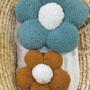 May include: Two flower-shaped decorative pillows. One is teal with a white center, and the other is brown with a white center. The pillows are made of a textured material and are placed in a woven basket.