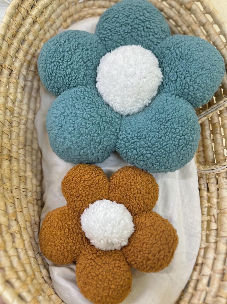 May include: Two flower-shaped decorative pillows. One is teal with a white center, and the other is brown with a white center. The pillows are made of a textured material and are placed in a woven basket.