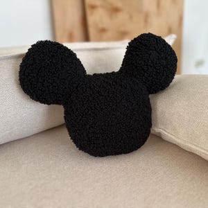 May include: A black, plush pillow in the shape of a cartoon mouse head with large, rounded ears. The pillow is made of a textured, fuzzy material and rests on a beige cushion. The ears are symmetrical and the overall design is simple and recognizable.
