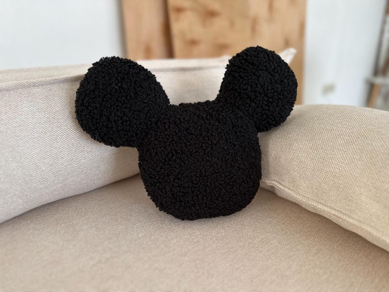 May include: A black, plush pillow in the shape of a cartoon mouse head with large, rounded ears. The pillow is made of a textured, fuzzy material and rests on a beige cushion. The ears are symmetrical and the overall design is simple and recognizable.