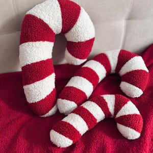 May include: Four plush candy cane decorations in red and white stripes. The candy canes are arranged on a red blanket and against a light-colored background. The soft, textured material gives a cozy feel, perfect for holiday decor.