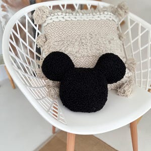 May include: A black, plush Mickey Mouse-shaped pillow rests on a white chair, with a beige patterned pillow behind it. The chair has a modern design with wooden legs. The background includes a neutral-toned rug and a glimpse of a plant.