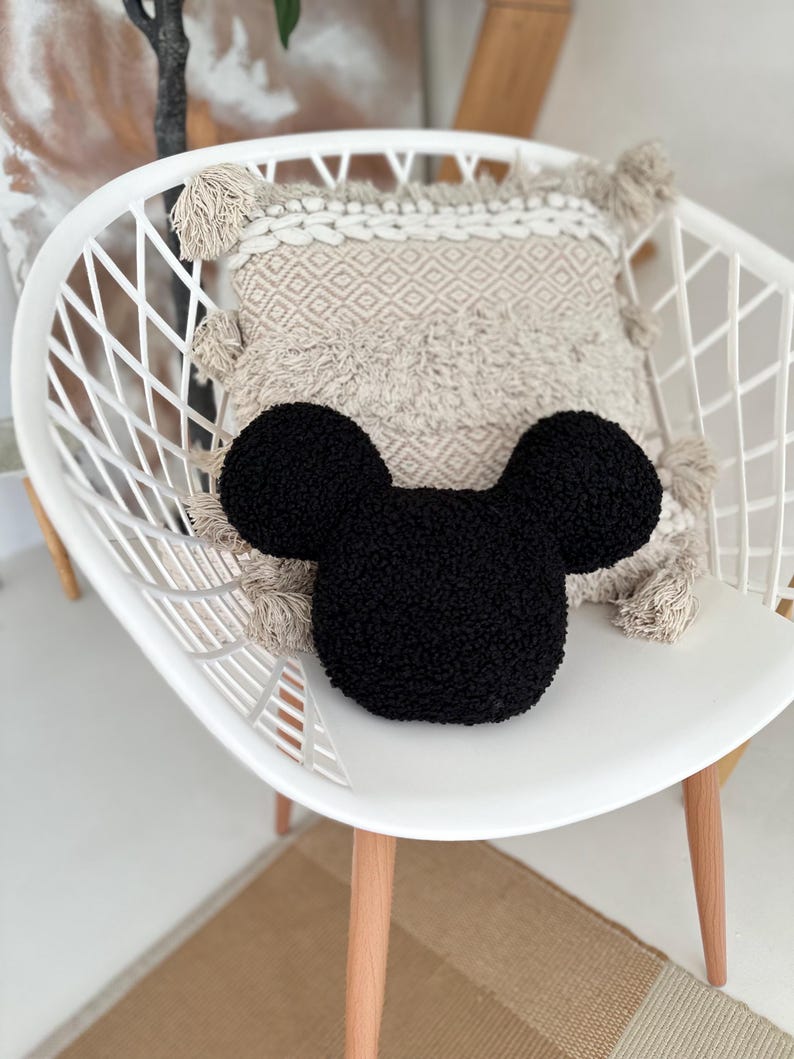 May include: A black, plush Mickey Mouse-shaped pillow rests on a white chair, with a beige patterned pillow behind it. The chair has a modern design with wooden legs. The background includes a neutral-toned rug and a glimpse of a plant.