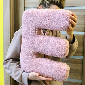 K&ouml;nnte beinhalten: Ein pl&uuml;schiges, buchstabenf&ouml;rmiges Kissen in Form des Buchstabens E, bezogen mit einem weichen, strukturierten, hellrosa Sherpa- oder Boucl&eacute;-Stoff. Das Kissen hat eine dicke, abgerundete Form und eine durchgehend flauschige Oberfl&auml;chenstruktur.