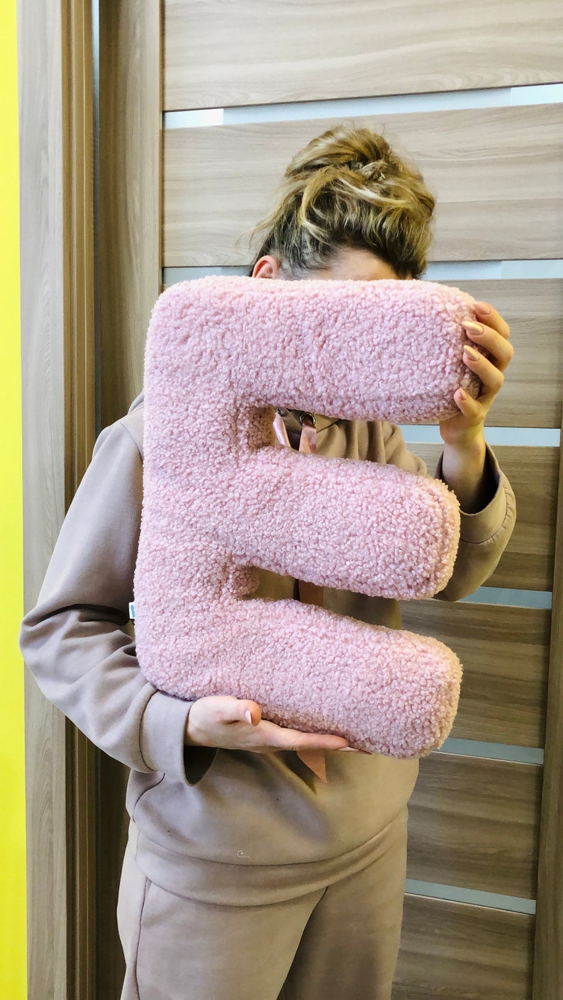 K&ouml;nnte beinhalten: Ein pl&uuml;schiges, buchstabenf&ouml;rmiges Kissen in Form des Buchstabens E, bezogen mit einem weichen, strukturierten, hellrosa Sherpa- oder Boucl&eacute;-Stoff. Das Kissen hat eine dicke, abgerundete Form und eine durchgehend flauschige Oberfl&auml;chenstruktur.