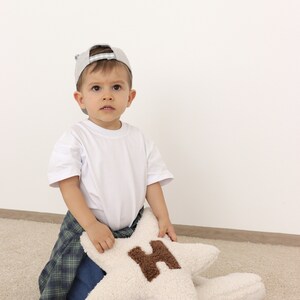 以下が含まれることがあります： グレーの野球帽と白いTシャツを着た幼い子供が、グレーと白の模様の敷物の上に座っています。子供は、茶色のHの文字が書かれた白い星型のクッションを持っています。