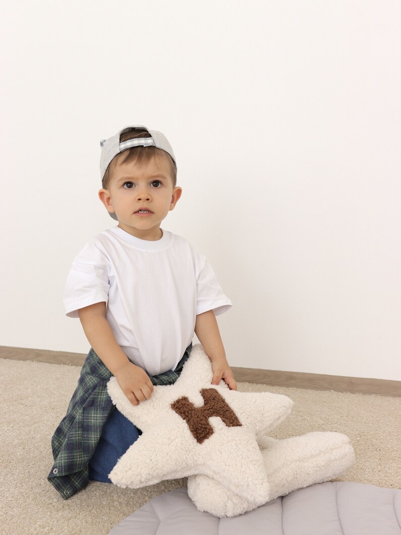 以下が含まれることがあります： グレーの野球帽と白いTシャツを着た幼い子供が、グレーと白の模様の敷物の上に座っています。子供は、茶色のHの文字が書かれた白い星型のクッションを持っています。