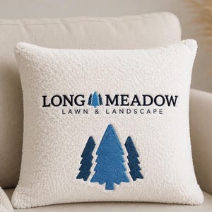 Może przedstawiać: Kwadratowa biała poduszka dekoracyjna typu sherpa z wyhaftowanym ciemnoniebieskim napisem LONG MEADOW LAWN & LANDSCAPE. Wzór zawiera małą niebieską ikonę drzewa między słowami oraz trzy większe niebieskie wyhaftowane sosny pod tekstem.