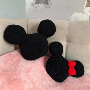 May include: Two black, plush Mickey Mouse and Minnie Mouse head-shaped pillows. The Mickey pillow has two large round ears. The Minnie pillow has a red bow and two round ears. The pillows are on a pink blanket.