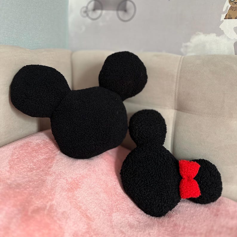 May include: Two black, plush Mickey Mouse and Minnie Mouse head-shaped pillows. The Mickey pillow has two large round ears. The Minnie pillow has a red bow and two round ears. The pillows are on a pink blanket.