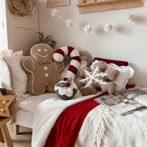 May include: A festive bedroom scene with Christmas-themed cushions. The cushions include a gingerbread man, a candy cane, a snowflake, and a Christmas pudding. A red throw blanket adds a festive touch to the white bedding.