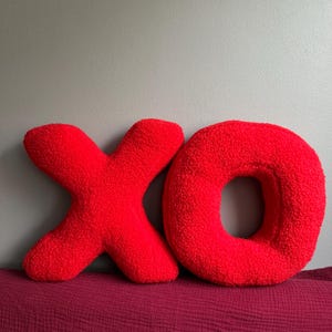 K&ouml;nnte beinhalten: Zwei leuchtend rote Dekokissen in Form der Buchstaben X und O. Die Kissen bestehen aus einem strukturierten, flauschigen, schafwollartigen Stoff. Der Buchstabe X befindet sich links neben dem kreisf&ouml;rmigen Buchstaben O und bildet das K&uuml;rzel XO.