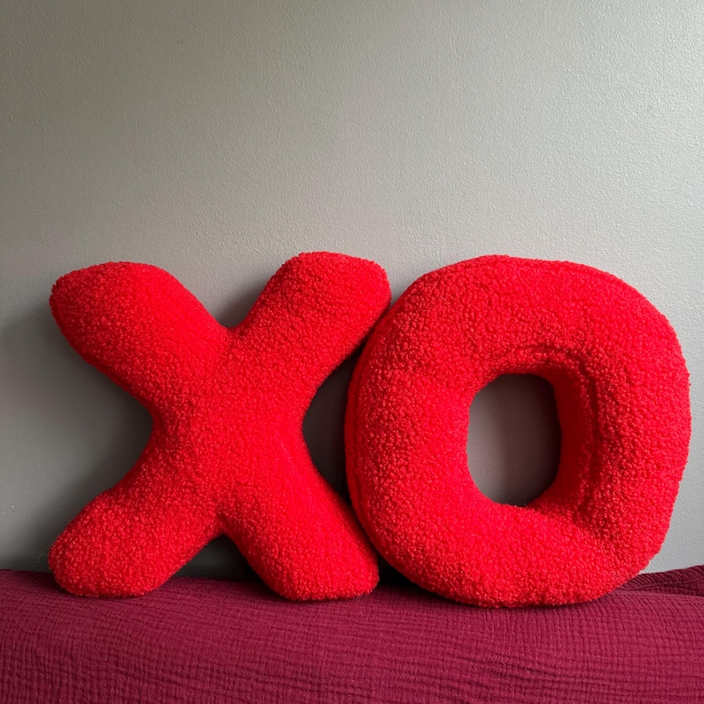 K&ouml;nnte beinhalten: Zwei leuchtend rote Dekokissen in Form der Buchstaben X und O. Die Kissen bestehen aus einem strukturierten, flauschigen, schafwollartigen Stoff. Der Buchstabe X befindet sich links neben dem kreisf&ouml;rmigen Buchstaben O und bildet das K&uuml;rzel XO.