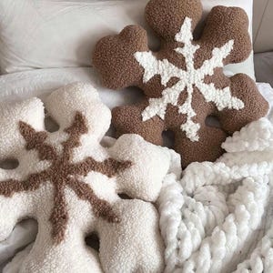 May include: Two plush snowflake-shaped pillows, one brown with a white snowflake design and one white with a brown snowflake design. The pillows are on a white fluffy blanket.