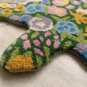 May include: Close-up of a floral-patterned rug in a star shape. The rug features a green background with various flowers in shades of pink, blue, yellow, and purple. The rug is made of a soft, textured material.