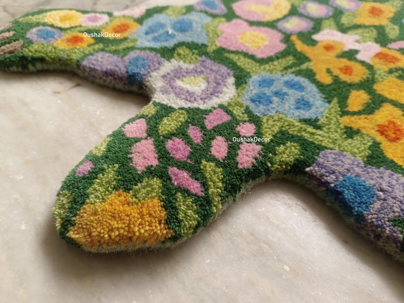 May include: Close-up of a floral-patterned rug in a star shape. The rug features a green background with various flowers in shades of pink, blue, yellow, and purple. The rug is made of a soft, textured material.