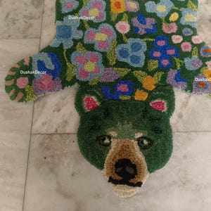 May include: A green bear-shaped rug with floral patterns. The rug features a bear head with a brown snout and black nose. The body of the rug is green with colorful flowers in shades of blue, pink, yellow, and orange. The brand name "OushakDecor" is visible.