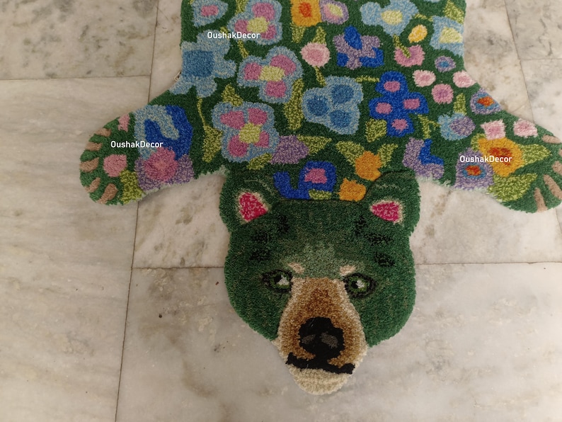May include: A green bear-shaped rug with floral patterns. The rug features a bear head with a brown snout and black nose. The body of the rug is green with colorful flowers in shades of blue, pink, yellow, and orange. The brand name "OushakDecor" is visible.