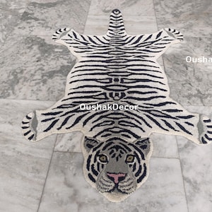 May include: A white and black tiger-shaped rug with a detailed face and striped pattern. The rug has a cream-colored background with black stripes and a gray face with pink accents. The rug is placed on a marble floor.