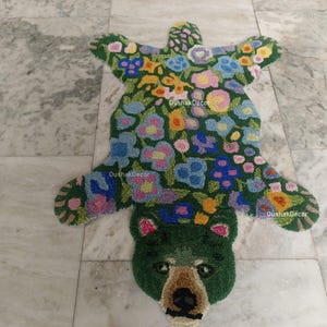 May include: A handmade rug in the shape of a bear, featuring a green base with colorful floral patterns. The bear's head is detailed with a brown nose and ears. The rug is placed on a marble floor.