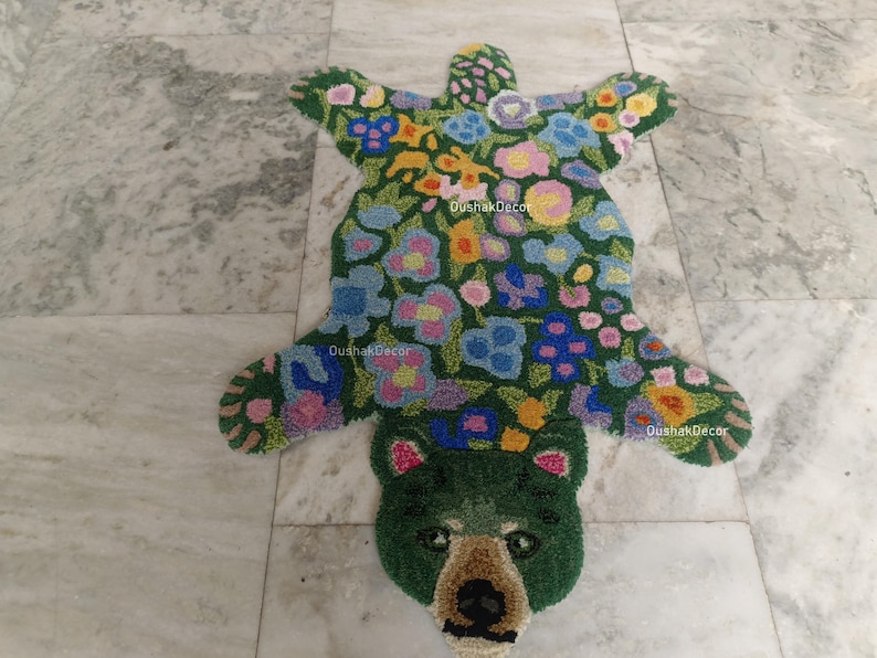 May include: A handmade rug in the shape of a bear, featuring a green base with colorful floral patterns. The bear's head is detailed with a brown nose and ears. The rug is placed on a marble floor.