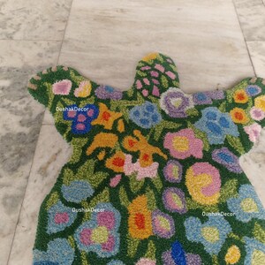 May include: A floral-patterned rug in a unique shape, featuring a green background with colorful flowers in shades of blue, yellow, pink, and purple. The rug has a textured appearance and the text "OushakDecor" is visible in several places.