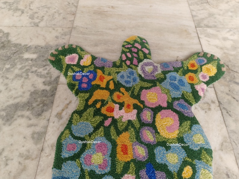 May include: A floral-patterned rug in a unique shape, featuring a green background with colorful flowers in shades of blue, yellow, pink, and purple. The rug has a textured appearance and the text "OushakDecor" is visible in several places.
