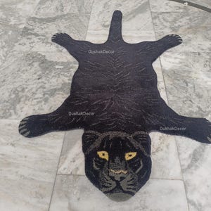May include: A black panther-shaped rug with yellow eyes and a detailed face. The rug is made of a dark material with a textured appearance, and the design includes the text "OushakDecor". The rug is laid out on a marble floor.