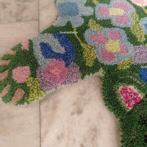 May include: Close-up of a colorful, floral-patterned rug. The rug features a green background with blue, pink, and purple flower designs. The rug is made of a textured material, and the design is detailed and intricate. The text "OushakDecor" is visible.