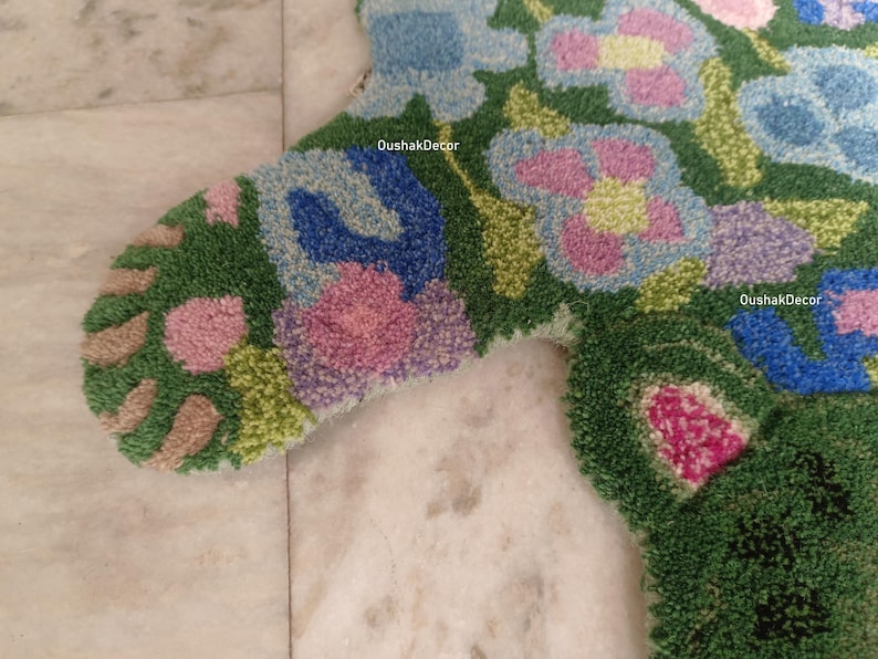 May include: Close-up of a colorful, floral-patterned rug. The rug features a green background with blue, pink, and purple flower designs. The rug is made of a textured material, and the design is detailed and intricate. The text "OushakDecor" is visible.