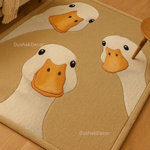 May include: A rectangular beige rug featuring three large, cartoon-style duck faces. The ducks have white heads, orange beaks, and black eyes. The rug has a brown border and the text "OushakDecor" is visible.