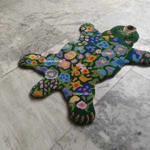 May include: A handmade, green bear-shaped rug with a floral design. The rug features a dark green background with colorful flowers in shades of pink, yellow, blue, and purple. The bear's face and paws are detailed with brown and green accents.