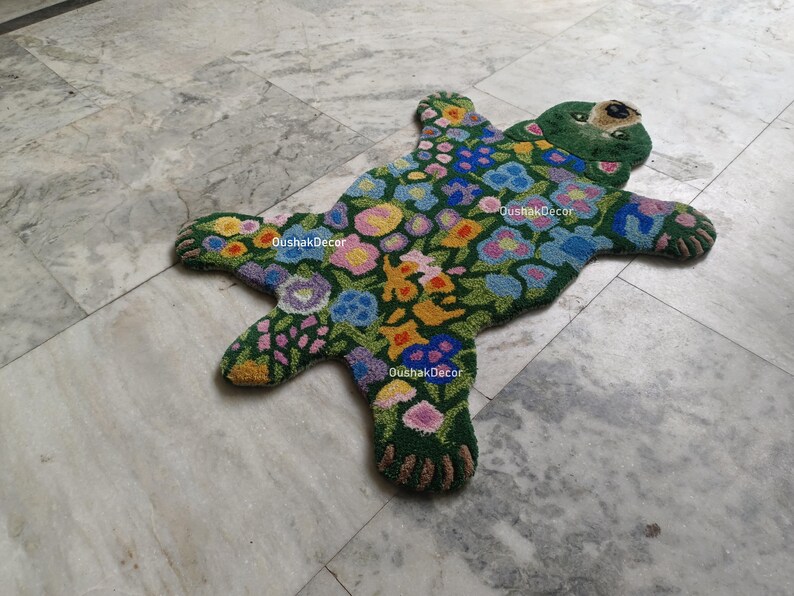 May include: A handmade, green bear-shaped rug with a floral design. The rug features a dark green background with colorful flowers in shades of pink, yellow, blue, and purple. The bear's face and paws are detailed with brown and green accents.