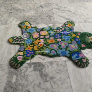 May include: A green rug in the shape of a bear, adorned with colorful floral patterns. The rug features a bear head with a brown nose and paws. The rug has the text "OushakDecor" in white.