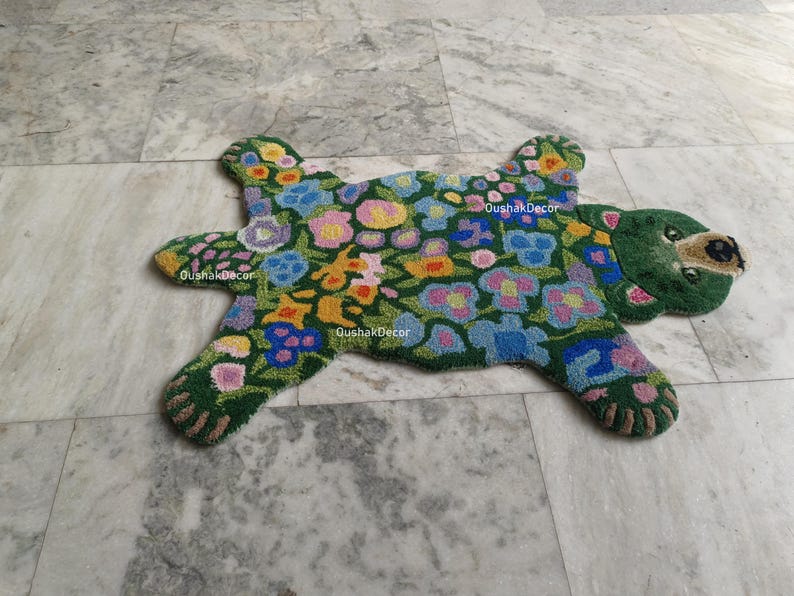 May include: A green rug in the shape of a bear, adorned with colorful floral patterns. The rug features a bear head with a brown nose and paws. The rug has the text "OushakDecor" in white.