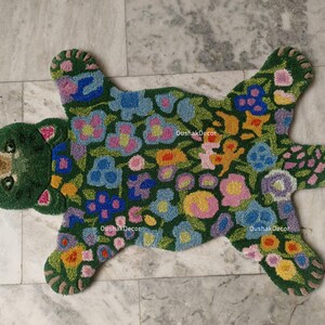 May include: A green bear-shaped rug with a floral pattern. The rug features a bear head and paws, with a body covered in colorful flowers. The rug is made of a soft, textured material and is designed for floor use.