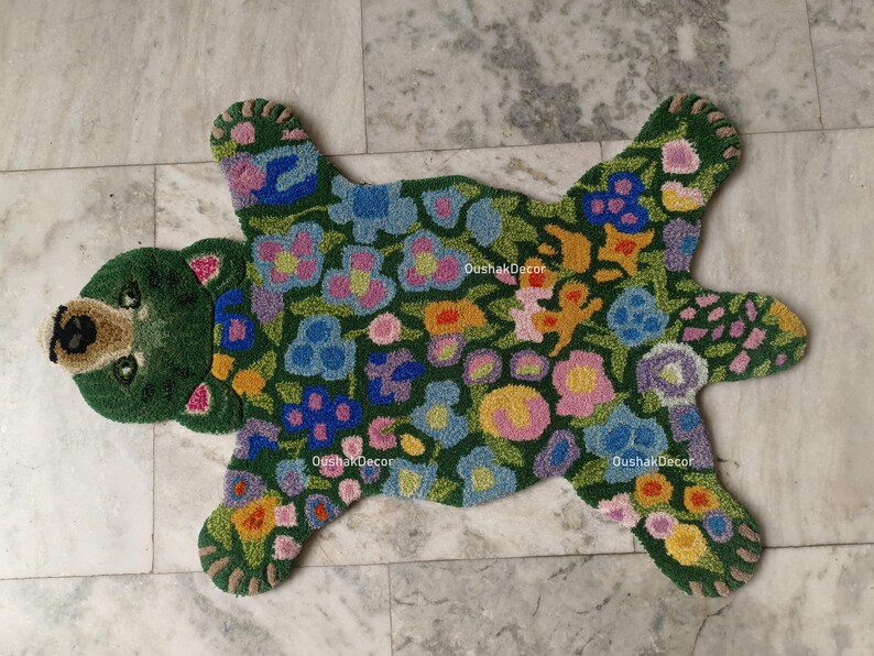May include: A green bear-shaped rug with a floral pattern. The rug features a bear head and paws, with a body covered in colorful flowers. The rug is made of a soft, textured material and is designed for floor use.