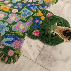May include: A green, floral-patterned rug in the shape of a bear. The rug features a green background with colorful flowers in shades of blue, pink, yellow, and orange. The bear's face is tan with black details. The rug is on a marble surface.