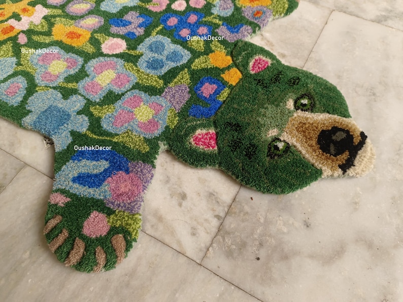 May include: A green, floral-patterned rug in the shape of a bear. The rug features a green background with colorful flowers in shades of blue, pink, yellow, and orange. The bear's face is tan with black details. The rug is on a marble surface.