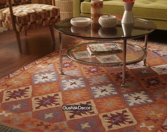 May include: An orange and brown patterned rug with a geometric design, fringed edges, and the text "OushakDecor". The rug is placed in a living room setting with a glass-topped coffee table, a green sofa, and a patterned armchair.