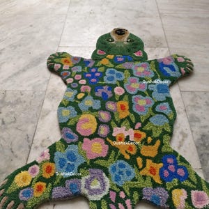 May include: A green, bear-shaped rug with a floral design. The rug features a green background with colorful flowers in shades of pink, blue, yellow, and purple. The bear's head is green with a brown nose and eyes.