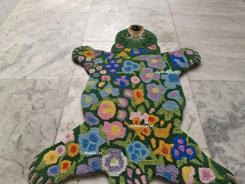 May include: A green, bear-shaped rug with a floral design. The rug features a green background with colorful flowers in shades of pink, blue, yellow, and purple. The bear's head is green with a brown nose and eyes.