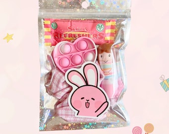Puede incluir: Una bolsa de plástico transparente llena de una pegatina de conejo rosa y blanco, un juguete pop-it rosa en forma de corazón, una muñeca a rayas rosa y blanca y una goma para el pelo a rayas rosa y blanca. La bolsa también contiene una barra de caramelo con el texto "Swizzels Refreshers" impreso en ella.