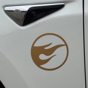 May include: A gold Hot Wheels logo sticker on a white car door. The logo is a flame design within a circle. A chrome side mirror is visible above the sticker.
