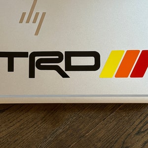 May include: A black and white "TRD" logo with three red, orange and yellow stripes.